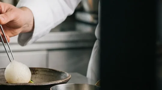 Ein Schweizer Spitzenkoch richtet in einer modernen Kueche ein kunstvolles Gericht auf einem handgefertigten Keramikteller an, im Hintergrund alpine Kraeuterbuendel